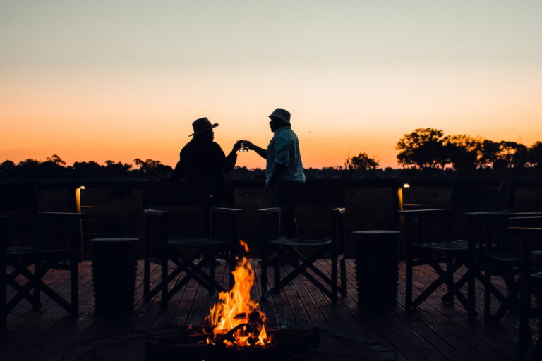wo guests sharing a drink around a campfire at sunset at Mma Dinare Camp.