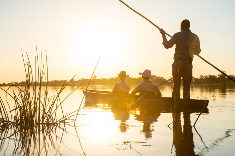 Guests in mokoro canoe on calm waters at sunset in the Okavango Delta near Kwara Camp.