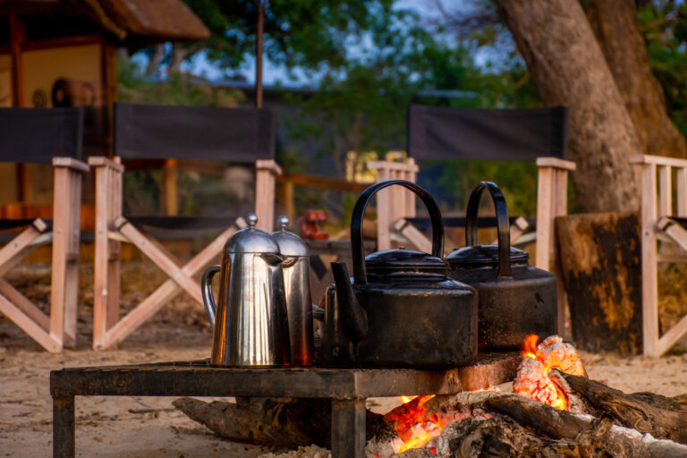 Coffee pots heating over an open campfire with chairs in the background.