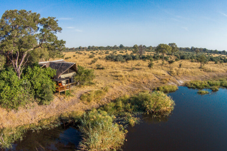 Aerial view of safari camp beside a lagoon with trees and golden grasslands.