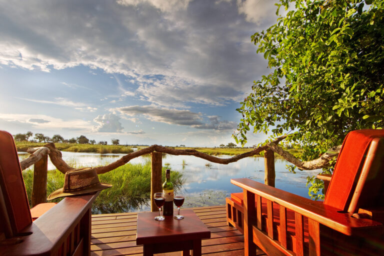 Two wooden chairs on a deck overlooking water with wine glasses on a table.