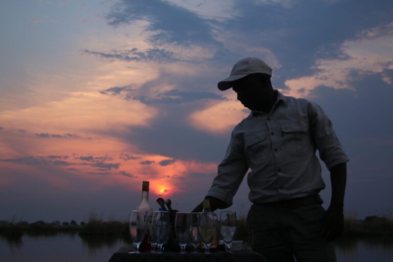 Two wooden chairs on a deck overlooking the river with two glasses of wine on a table.
