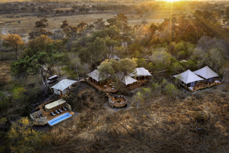 Aerial view of a luxury safari lodge surrounded by trees and savannah at sunset, with tents, a pool, and outdoor seating areas.
