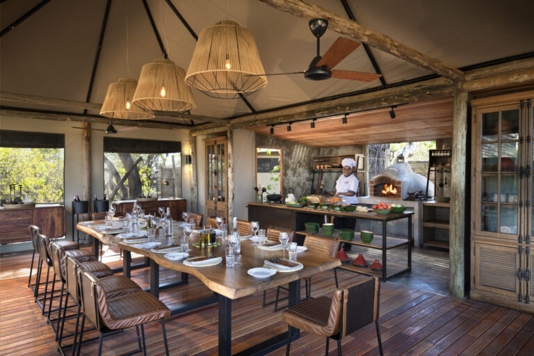 Elegant open-air dining area with a long wooden table and a chef preparing food in a rustic kitchen with a pizza oven.