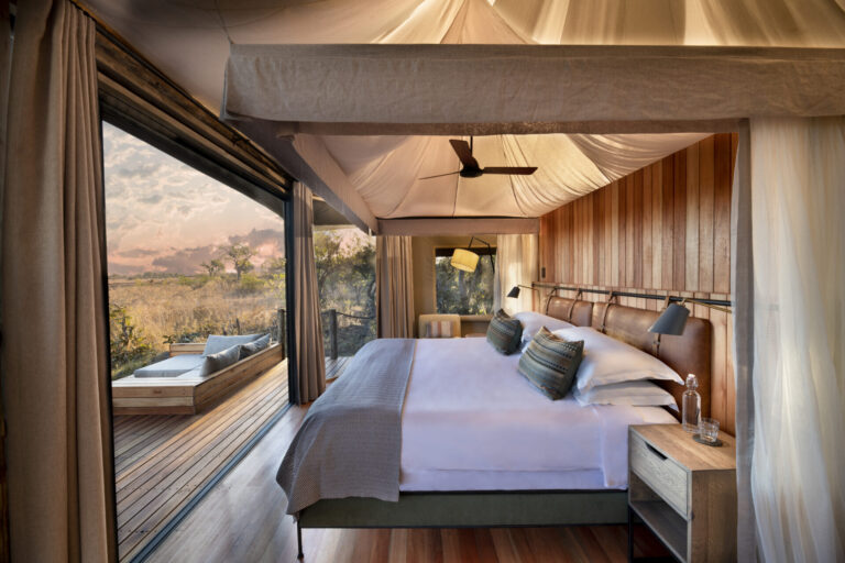 Spacious safari tent with a king-size bed, wooden interior, and sliding glass doors opening to a private deck overlooking the African bush.
