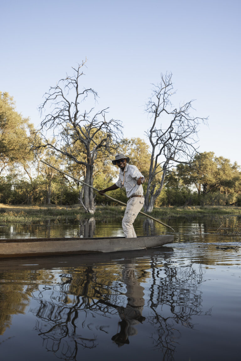 A guide in traditional attire steers a mokoro canoe through calm waters, surrounded by trees and reflections.