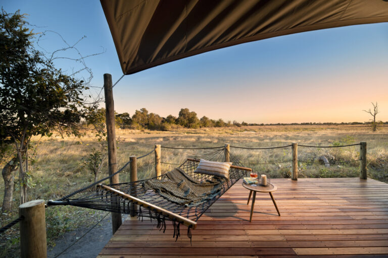 A hammock with pillows on a wooden deck overlooking a vast savannah under a clear sunset sky.