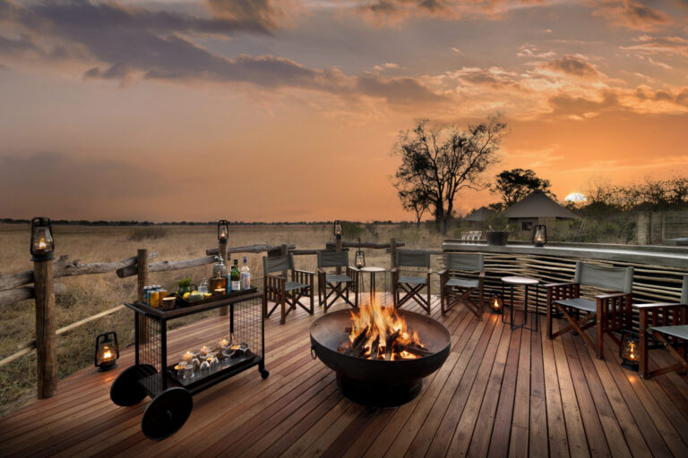 Wooden deck with fire pit, safari chairs, and drink cart overlooking the savannah at sunset.