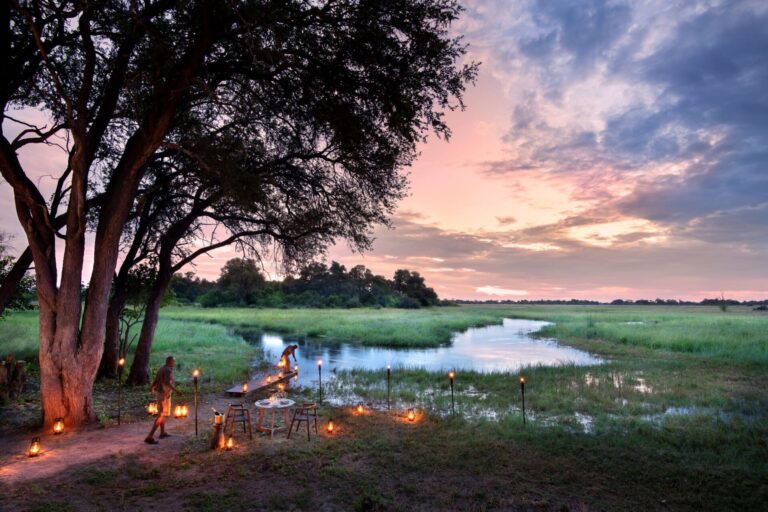Romantic outdoor dinner setup beside a tranquil river in the savannah, illuminated by lanterns at sunset.