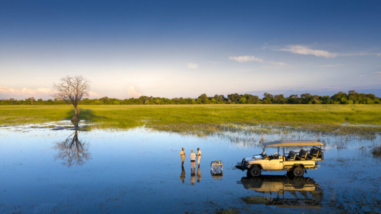 Three people stand in shallow water beside a safari vehicle enjoying drinks in the Okavango Delta at sunset.