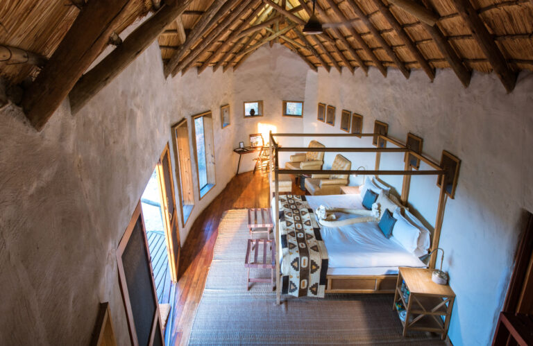 Aerial interior view of thatched chalet with a large bed and writing desk at Tau Pan Camp.