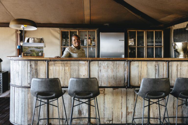 Bartender behind wooden safari bar at Linyanti Bush Camp in Botswana.